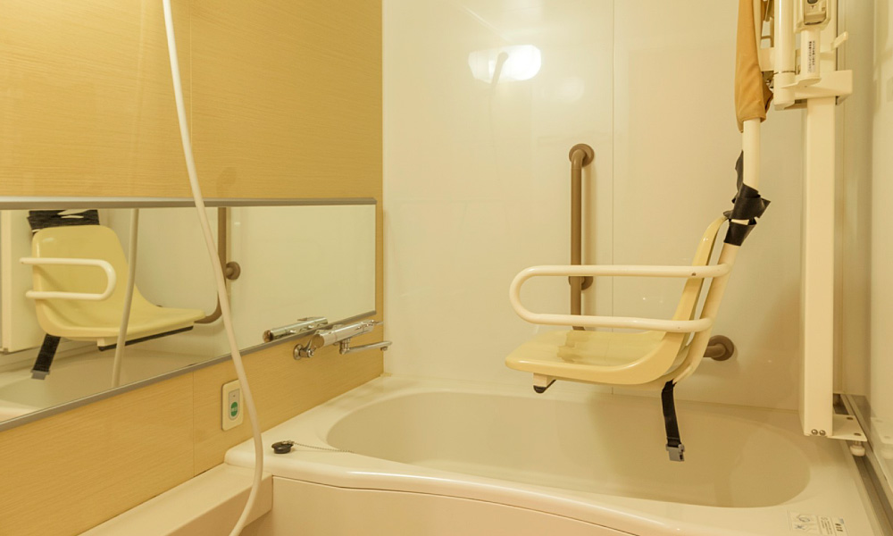 介護用リフト付きのバリアフリー浴室。浴槽の上に肘掛けとベルト付きの昇降いすが設置され、壁には縦手すりと横長の鏡、ハンドシャワーが備え付けられている。車椅子利用者なども安全に入浴しやすい設備。