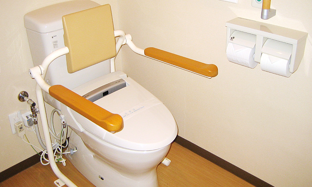 背もたれ付きの洋式トイレに、左右に長い肘掛け手すりを備えたバリアフリートイレ。立ち座りを補助するための手すりと温水洗浄便座、壁にはトイレットペーパーが二連で設置されている。