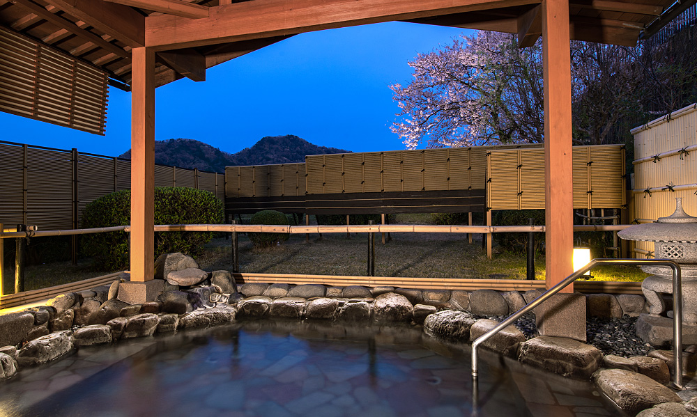 夕暮れの露天風呂。岩で縁取られた湯船の周囲を木の手すりと柱が囲み、奥には竹垣と庭木、咲き始めた桜と山並みが見える落ち着いた和風の景色。