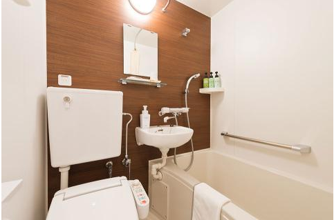 コンパクトなバスルーム。白い壁に木目調のアクセントがあり、洗面台、トイレ、バスタブが一体となっている。バスタブには手すりが設置されている。