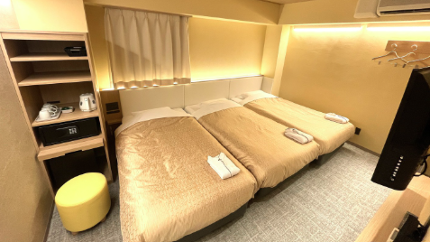  3つのシングルベッドが並ぶコンパクトなホテルルーム。ベッドにはそれぞれタオルが置かれ、黄色の壁が明るい雰囲気を演出している。棚にはティーセットとセーフティボックスがある。
