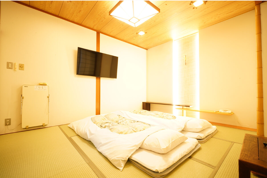 こちらは、和室の寝室です。畳敷きの和室に布団が敷かれており、壁にはテレビが取り付けられている。柔らかい照明が部屋を照らし、伝統的な日本の雰囲気を感じさせる。