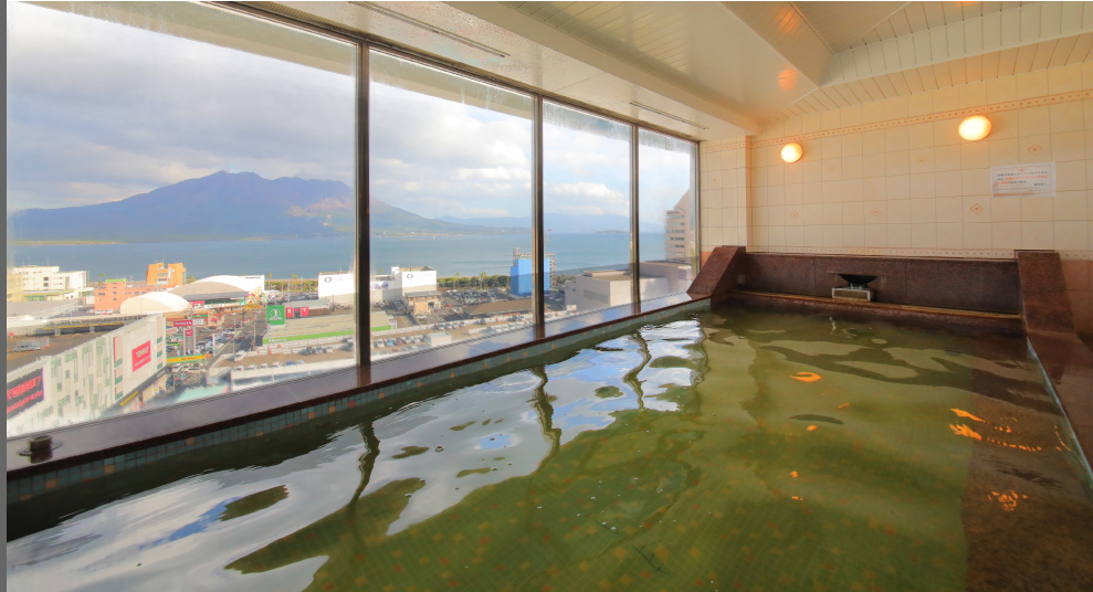 ホテルの展望風呂です。大きな窓からは街並みと海、遠くの山々が見渡せる。浴槽の水面が光を反射しています。
