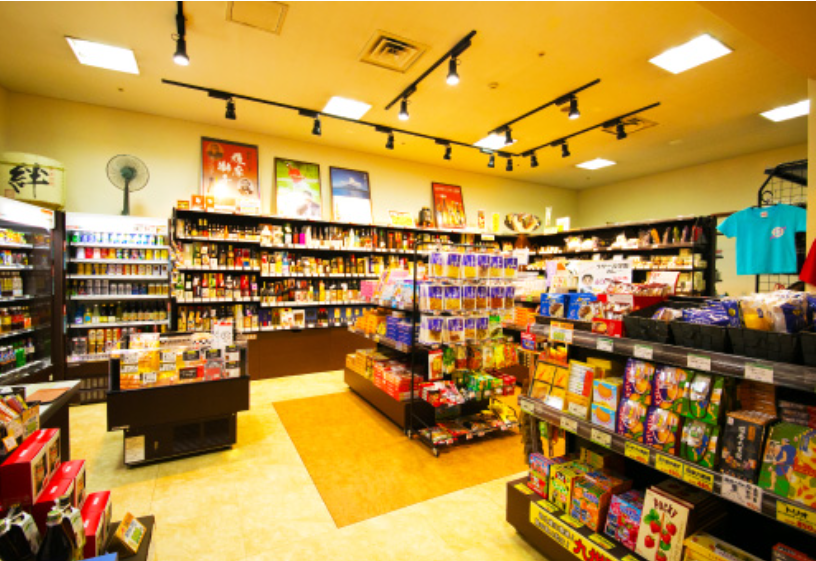 明るく照らされたホテルの売店です。棚には多くの飲み物やお菓子、土産物が並んでおり、中央には商品がディスプレイされています。