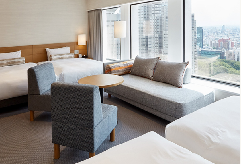 都市の景色を見渡せる大きな窓のあるホテルルーム。部屋には3つのベッド、グレーのソファ、丸いテーブル、2つの椅子が配置されている。