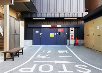 駐車場の入り口。地面には「止まれ」と「STOP」の表示があり、正面には青いシャッターが閉まっている。右側には駐車券の発券機と赤いコーンが置かれている。左側には階段とベンチが見える。