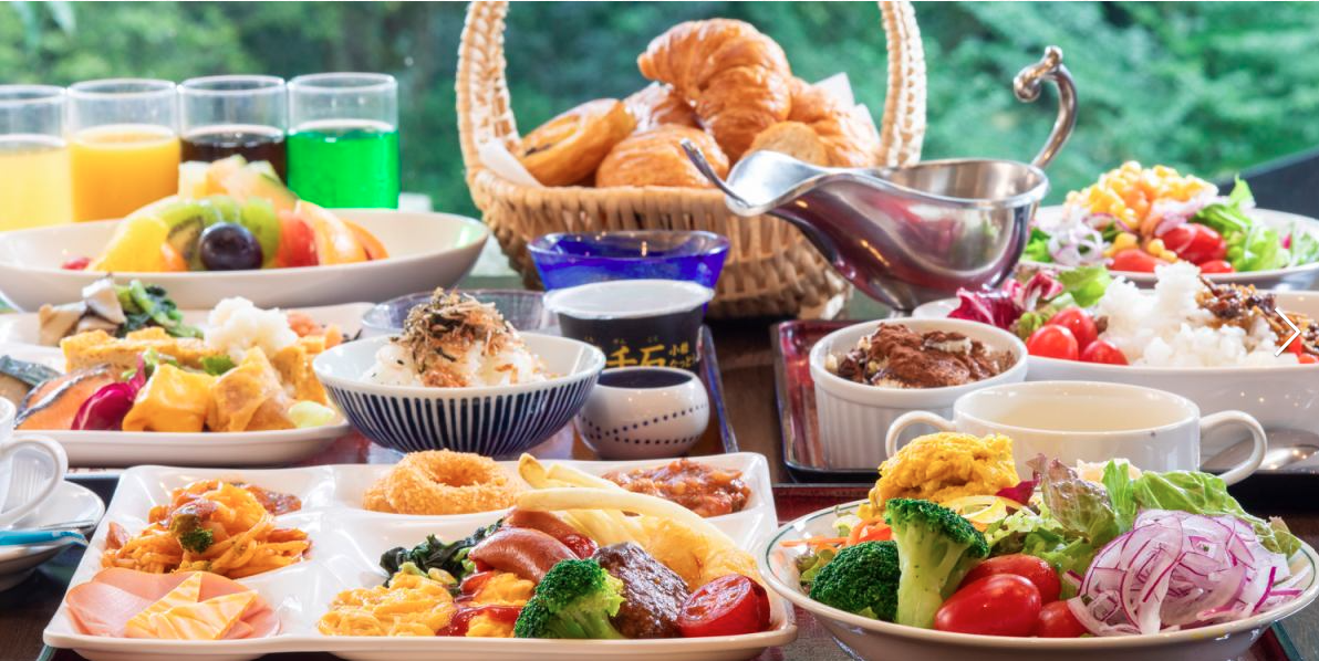 色とりどりの料理が並ぶビュッフェスタイルの朝食。フルーツ、サラダ、パン、ジュースなどがテーブルいっぱいに並べられ、窓の外には緑の景色が広がっている。