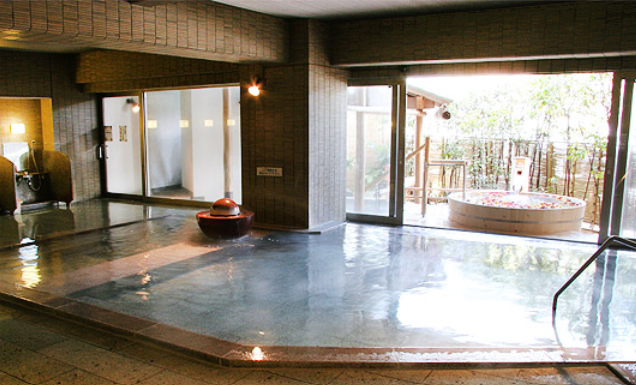こちらは、屋内温泉の画像です。大きな窓に面した屋内温泉。湯船の中央には陶器の装飾があり、外には小さな露天風呂が見えます。温泉の周囲にはタイルが敷かれ、自然光が差し込んでいます。