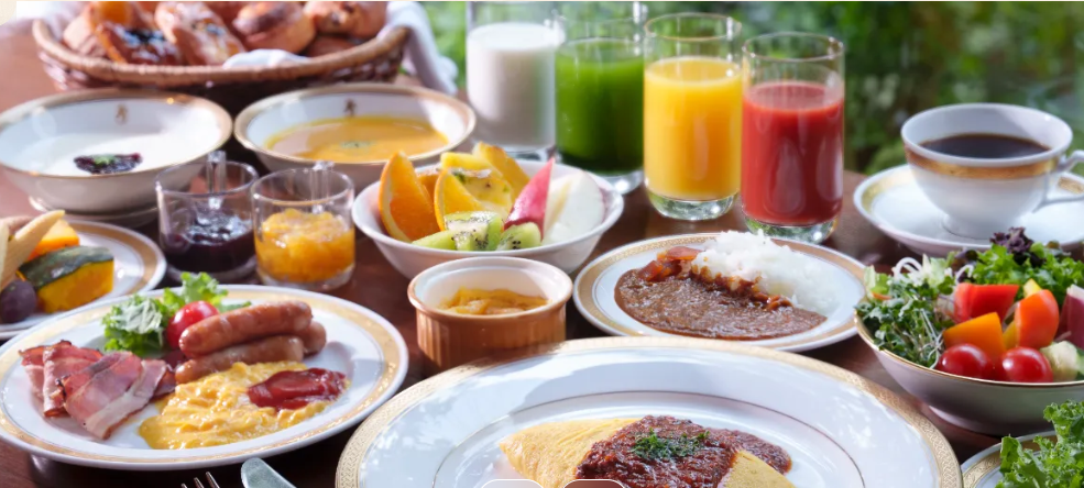色とりどりの朝食のテーブル。オムレツ、ベーコン、ソーセージ、カレーライス、サラダ、フルーツ、パンが並んでいる。飲み物として、緑、オレンジ、赤のジュースとコーヒーがある。全体的に豊富なバリエーションの料理が並び、朝食の楽しさを感じさせる。