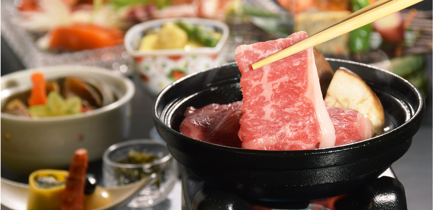 和風の食事シーンです。鉄板で焼かれている厚切りの和牛肉が箸で持ち上げられ、背景には色とりどりの料理が並んでいます。