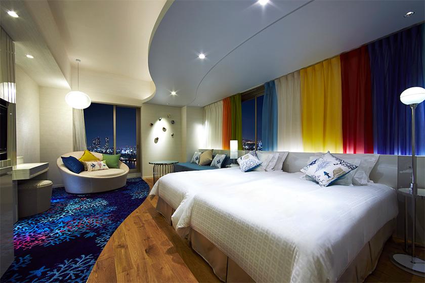夜のホテルのツインベッドルームの写真です。部屋は落ち着いた照明で照らされており、壁には赤、黄色、青のカーテンが掛けられています。ベッドには白いシーツと花柄のクッションが並べられています。窓からは夜景が見え、左側には円形のソファとテーブルが配置されています。