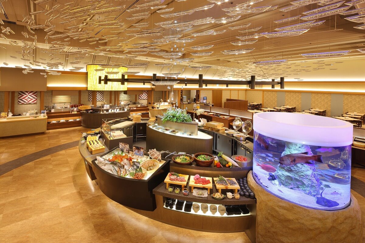 豪華なビュッフェレストランの内部です。中央には色とりどりの料理が並べられ、右側には大きな水槽があり、魚が泳いでいます。