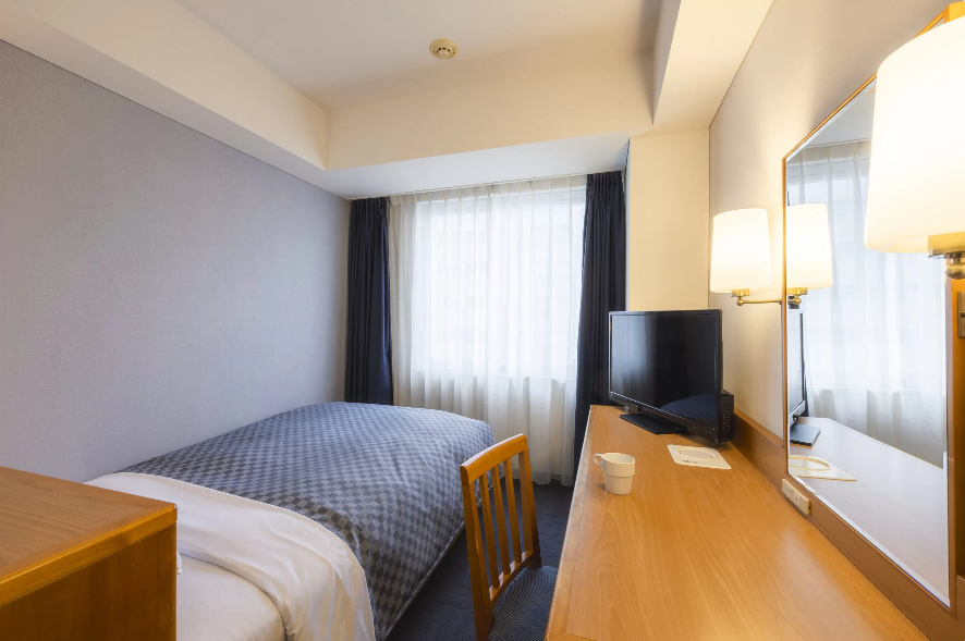 ホテルのシングルルームの写真。部屋には1台のベッドがあり、白いシーツとグレーのチェック柄カバーが掛けられている。ベッドの右側にはデスクがあり、上にはテレビとデスクライトが配置されている。デスクには椅子が置かれ、作業や食事に利用できる。右側には大きな窓があり、自然光が部屋に差し込んでいる。
