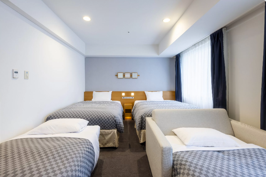 ホテルのファミリールームの写真。部屋には4台のベッドが並んでおり、白いシーツとグレーのチェック柄カバーが掛けられている。ベッドの間には小さなナイトスタンドがあり、壁には装飾的なアートが飾られている。右側には大きな窓があり、自然光が部屋に差し込んでいる。