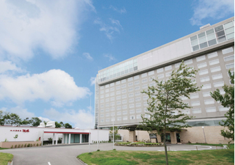 青空の下に立つ大きな白い近代的な建物。手前に緑の芝生と若木が植えられている。建物は複数階建てで、正面には大きな窓が並んでいる。建物の左側には低層の別棟が見える。