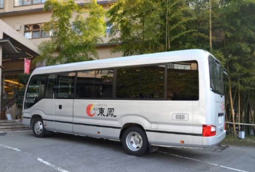 シルバーの小型バスが建物の前に停車している。バスの側面には「東風」と書かれたロゴがあり、ロゴには赤とオレンジの曲線が描かれている。背景には竹が植えられており、建物の窓が見える。