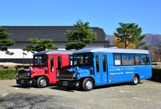 クラシックなデザインの赤と青のバスが並んで駐車されている。青いバスの側面には「まちなか周遊バス ハイカラさん」と書かれており、「Classic Town Bus Hakone-san」とも記載されている。背景には松の木と大きな建物があり、遠くには山が見える。