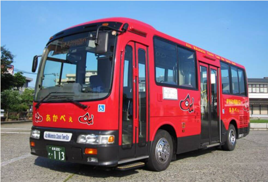 赤いボディに「あかべぇ」と書かれたイラストが描かれたバスが駐車場に停車している。バスの前面には「Akabeko Bus Service」と表示されており、ナンバープレートには「113」と記載されている。バスのドアには車椅子マークがあり、アクセシビリティに配慮されていることが示されている。背景には青空と建物が見える。