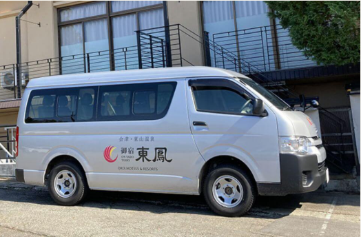 シルバーのバンが建物の前に停車している。バンの側面には「御宿 東鳳」と書かれたロゴがあり、赤とオレンジの曲線が描かれている。ロゴの下には「ONSEN HOTELS & RESORTS」と記載されている。背景には窓のある建物があり、階段が見える。バンは宿泊施設の送迎車のようです。