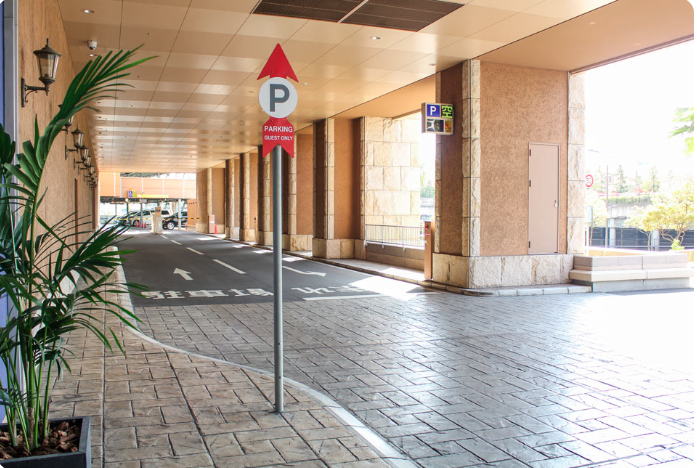 ホテルの駐車場入り口の写真。広い通路があり、右側には駐車場の標識が掲示されています。標識には赤い矢印と「P」の文字が描かれ、下に「PARKING GUEST ONLY」（宿泊者専用駐車場）と記載されています。通路の両側には石造りの壁が並び、天井は淡い色で統一されています。左側には植木鉢に植えられた観葉植物があり、通路の奥には駐車場が続いています。駐車場の入り口には「ENTER」（入口）と書かれた文字が路面に描かれています。