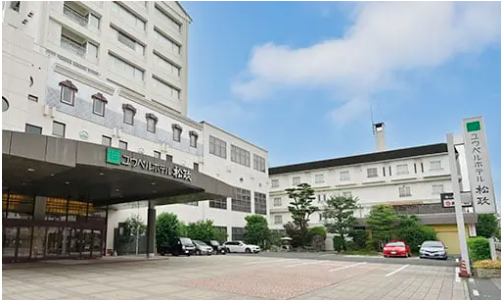 この画像は湯田温泉ユウベルホテル松政の外観を示しています。ホテルの正面には「ユウベルホテル松政」と書かれた看板があり、エントランスはガラス扉で構成されています。建物は白を基調としたデザインで、複数階建てです。エントランス前には広い駐車スペースがあり、数台の車が停まっています。周囲には緑の植栽が施され、青空が広がる晴れた日です。この画像はホテルの雰囲気やアクセスの様子を伝えるものです。