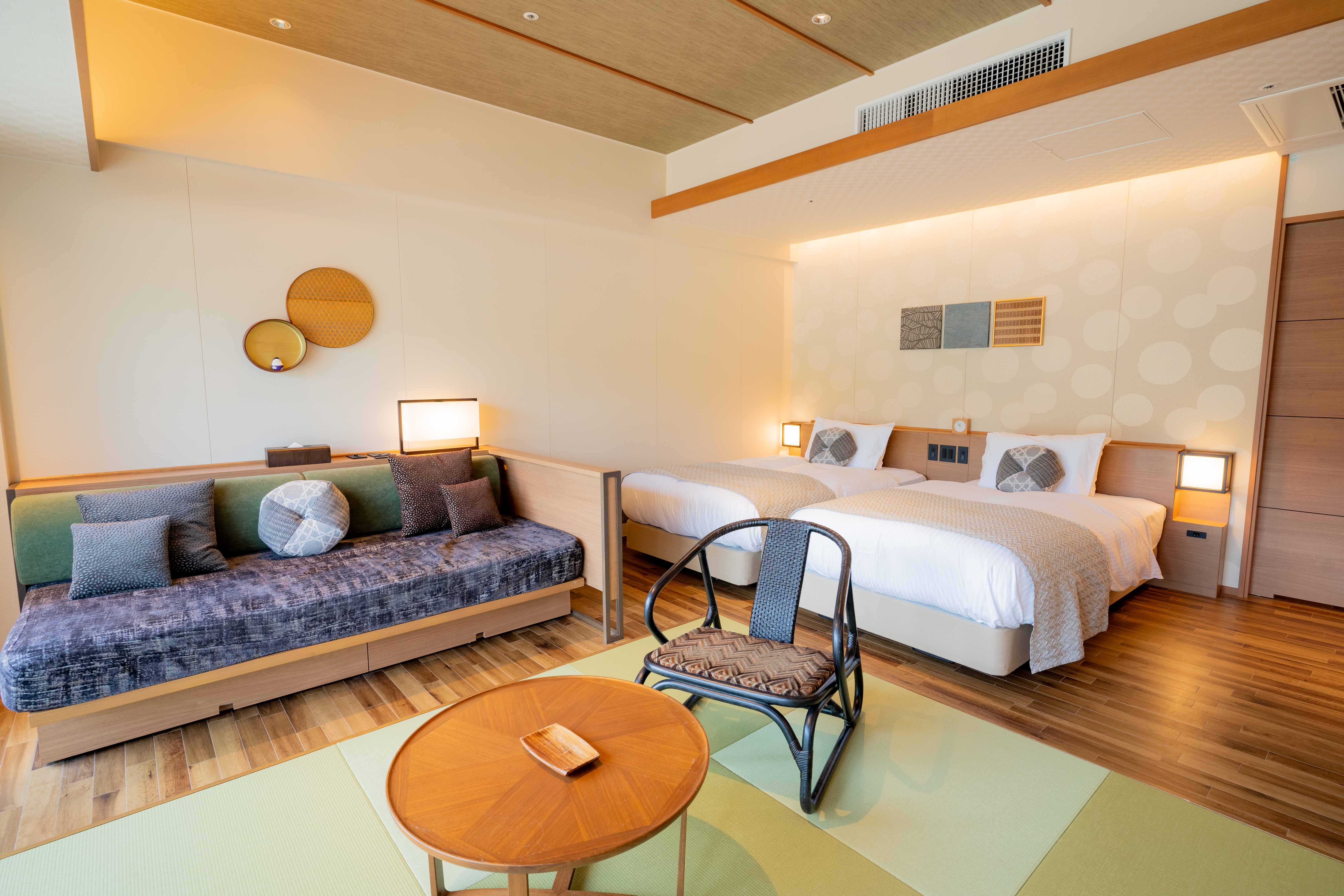 白いリネンと模様入りの枕が置かれたシングルベッドが2台並ぶ、モダンで明るいホテルの客室。ベッドの横にはカラフルなクッションが置かれたソファと、丸い木製テーブル、椅子がある。木製の床と円形模様の壁、ベッド上の装飾アート、複数の照明が室内を温かく照らしている。
