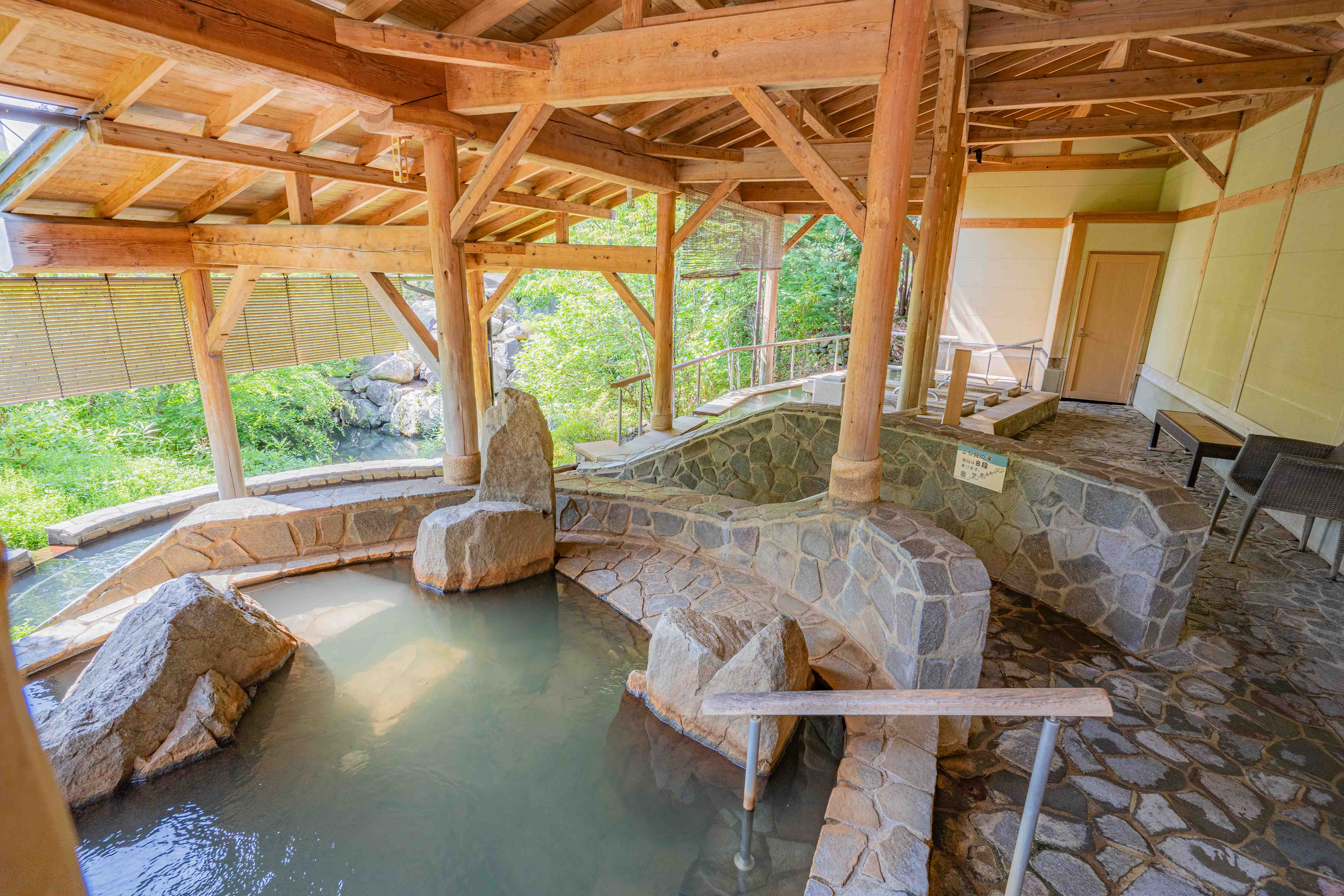 木造の屋根と石造りの壁に囲まれた、自然の緑に包まれた露天温泉。湯船には大きな岩が配置されており、奥には石壁に小さな看板と、扉へと続く小道が見える。静かで落ち着いた雰囲気の癒しの空間。
