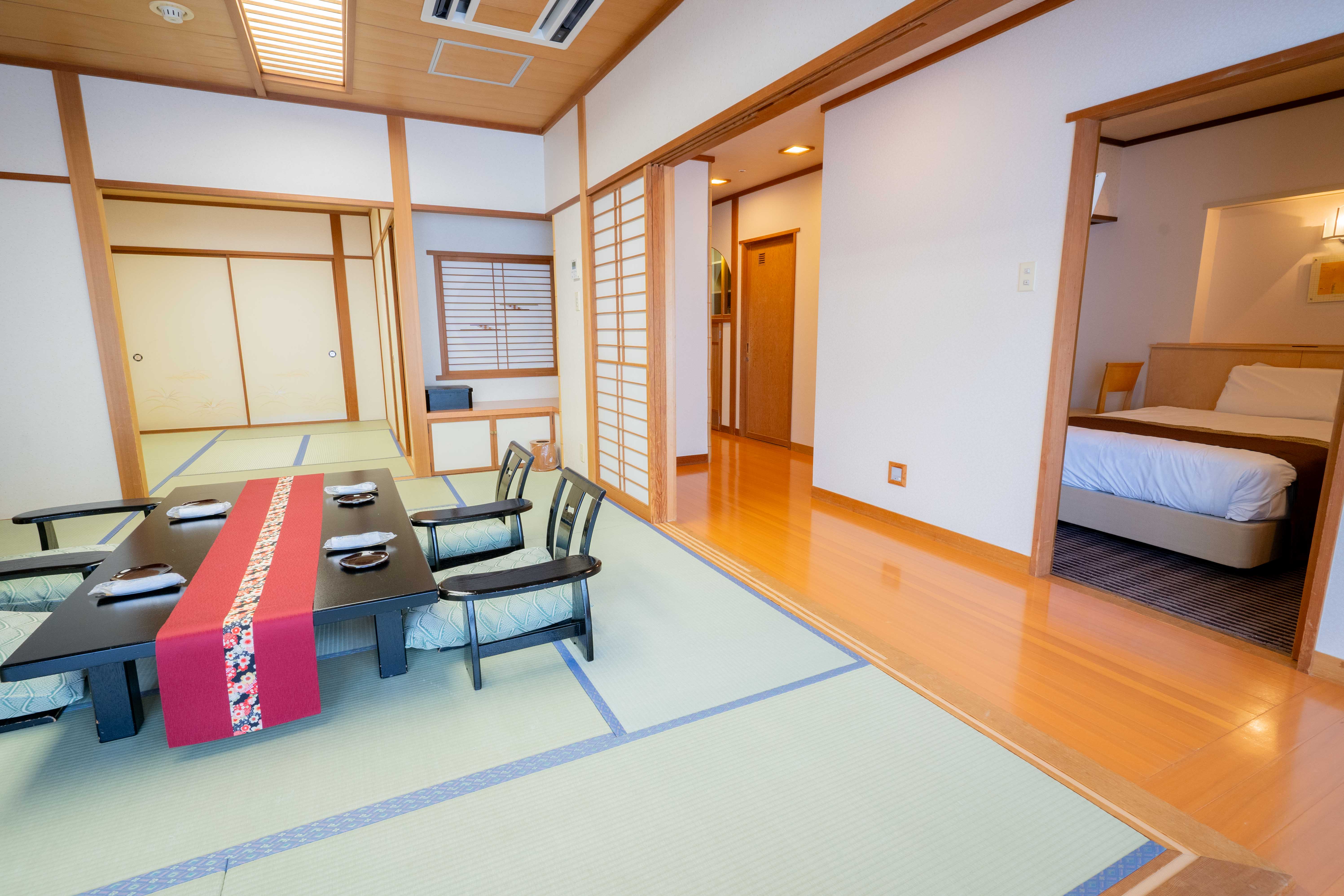 畳敷きの和室に、赤いテーブルランナーを敷いた黒い低めのテーブルと座布団付きの椅子が4脚並ぶ。障子と木の柱が和の雰囲気を演出し、奥の開いた戸の向こうには洋式ベッドが置かれた部屋が見える。和と洋が調和した清潔感のある室内。