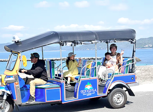 青い三輪タクシー（トゥクトゥク）が海岸沿いの道路を走行中。運転手と4人の乗客が乗っており、乗客は楽しそうに手を振っている。背景には青い海と山が広がっている。