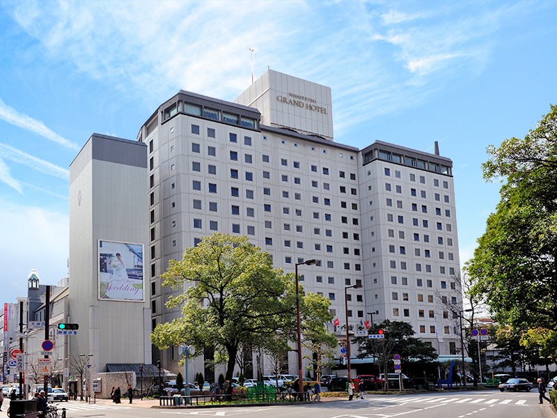 和晴天の下、交差点に面した『GRAND HOTEL』の白い建物洋折衷の客室。手前にはダイニングテーブルと椅子があり、奥にはツインベッドが配置されている。障子が部屋を仕切り、天井には丸い照明が吊るされている。