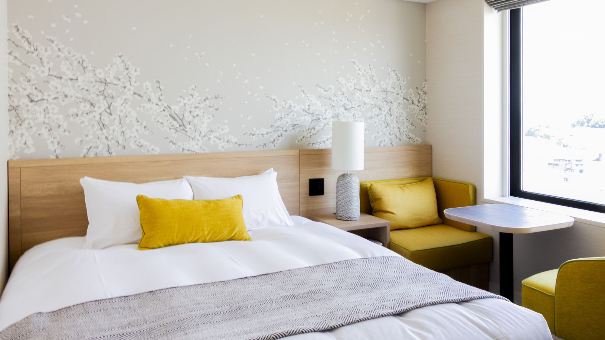 白いリネンと黄色のアクセントピローが置かれた大きなベッドがあるモダンなホテルの客室。ベッドの背面には白い花が咲く枝の装飾が施された壁があり、右側には黄色の椅子2脚と木製テーブルのある小さなシーティングエリアが、大きな窓のそばに配置されている。ベッドと椅子の間には白い円筒形のランプが置かれたナイトテーブルがある。