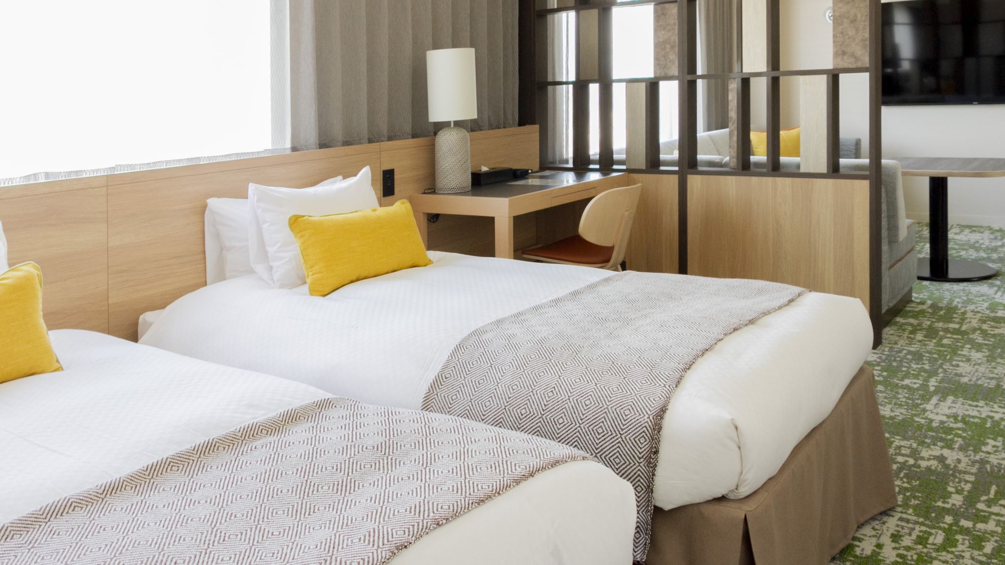 白いリネンとベージュの柄入りスロー、黄色のアクセントピローが置かれたシングルベッドが2台並ぶホテルの客室。ベッドの間には木製のヘッドボードがあり、デスクスペースとして延長され、椅子とランプ、装飾品が置かれている。緑の模様入りカーペットとナチュラルな色調の家具が特徴で、大きな窓から自然光が差し込む。