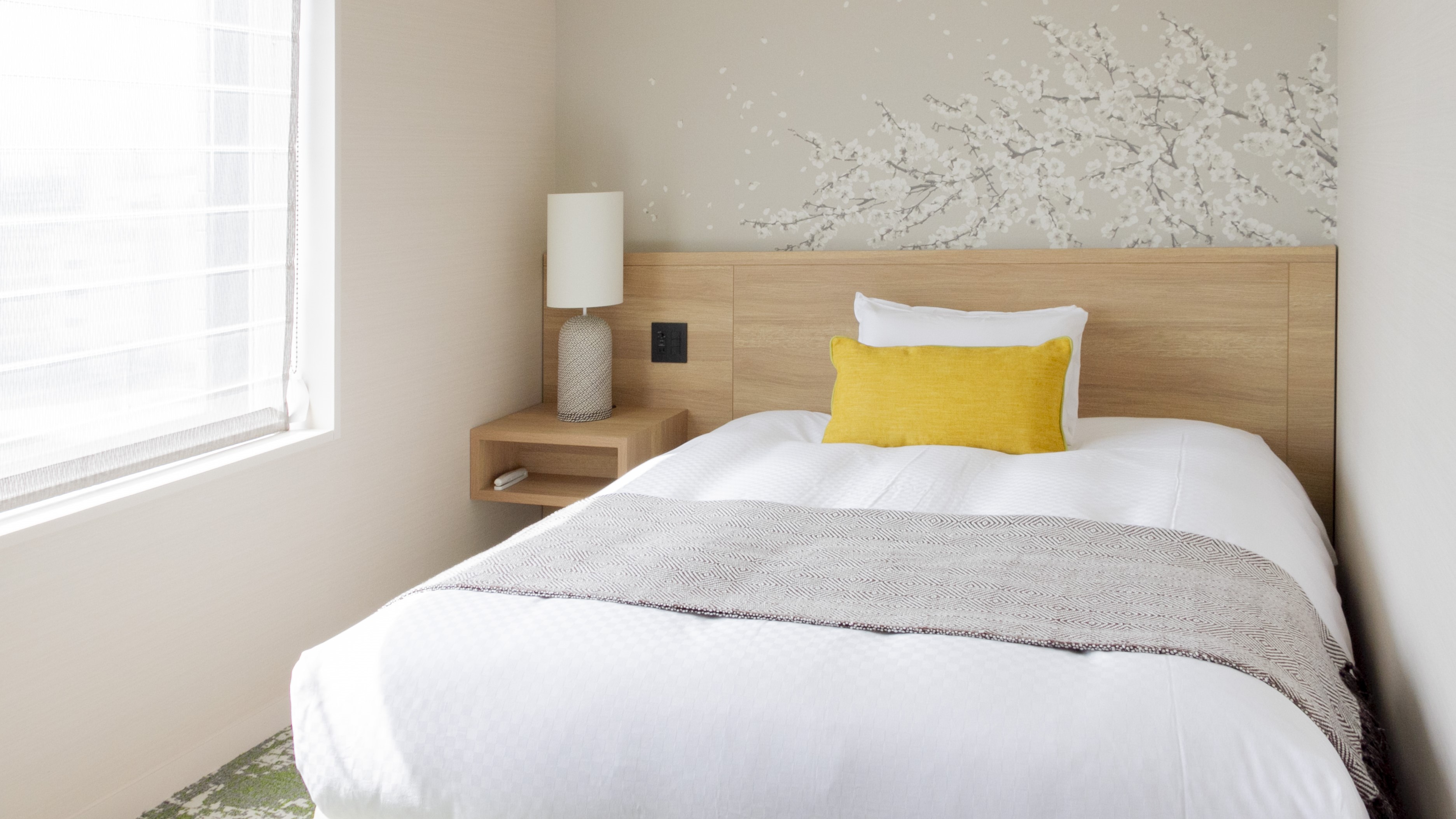 白いリネンとグレーのスロー、黄色のアクセントピローが置かれたベッドがあるミニマルな寝室。ベッドの左側には、白い円筒形のランプが置かれた木製のナイトスタンドがあり、壁には白い花が咲く枝の装飾が施されている。左側の大きな窓から自然光が差し込んでいる。