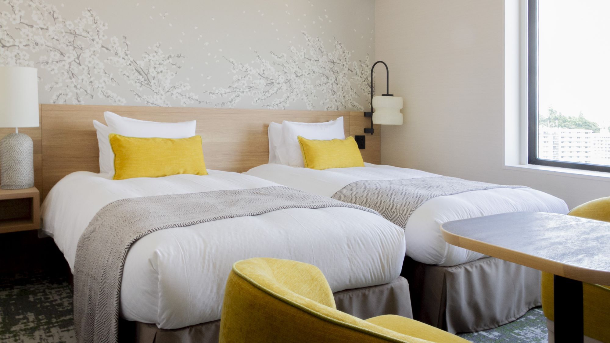 白いリネンとグレーのスロー、黄色のアクセントピローが置かれたシングルベッドが2台並ぶホテルの客室。木製のヘッドボードの背後には白い花が咲く枝の装飾が施された壁があり、左側にはランプ付きのナイトスタンドがある。右側の窓から自然光が差し込み、ベッドの前には黄色の椅子2脚と小さなテーブルが配置されている。