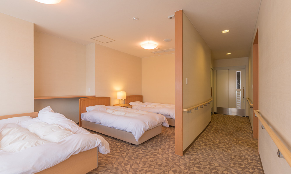バリアフリー対応の洋室。手前にシングルベッド1台、奥にシングルベッド2台が並び、廊下には両側に手すりが設置されている。入口まで段差が少なく、車椅子でも移動しやすい造りの明るい客室。