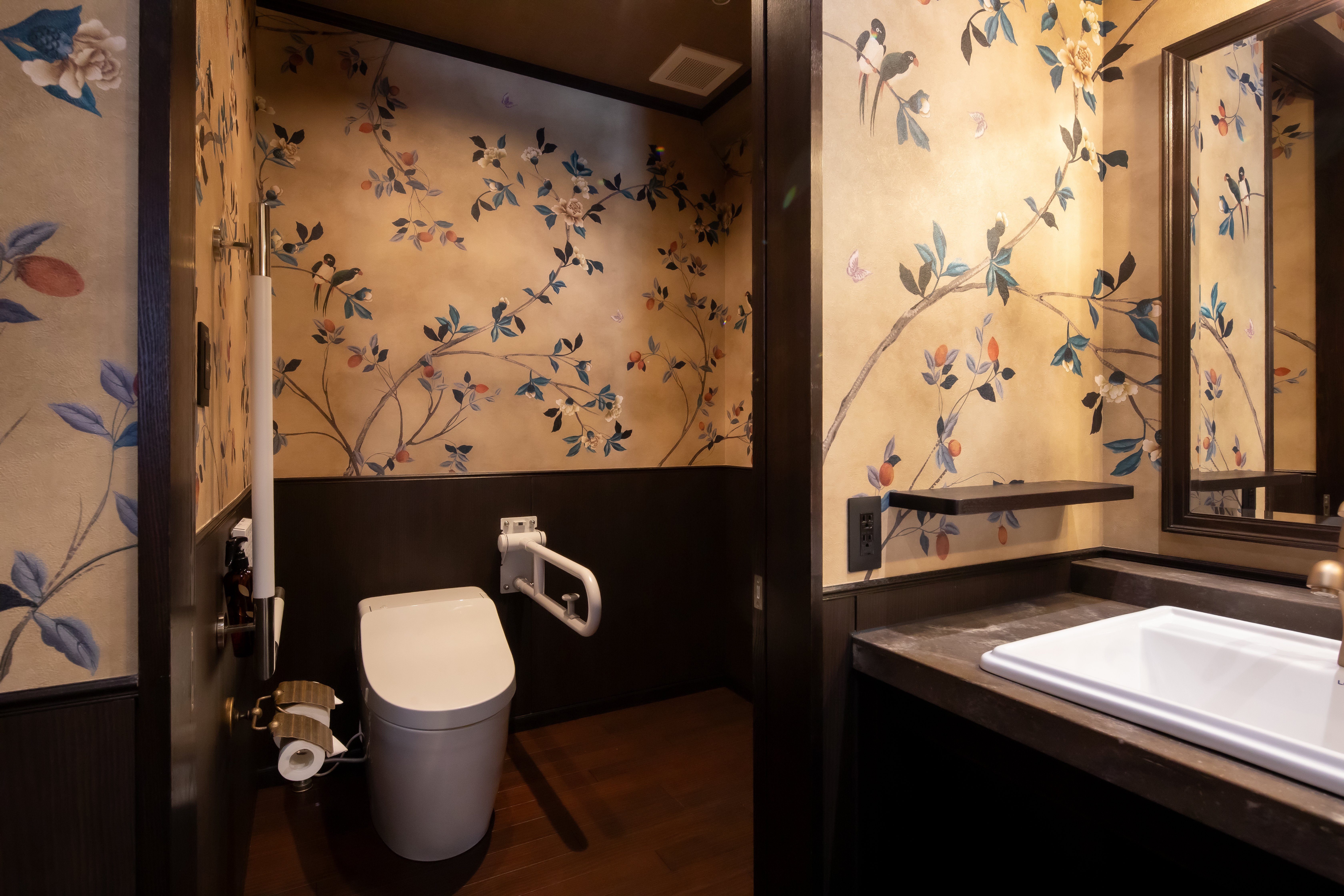 折りたたみ式手すりを備えた多目的トイレの内部。花と鳥の絵柄の壁紙が施された和モダンな内装で、洋式便器・洗面台・鏡を設置。