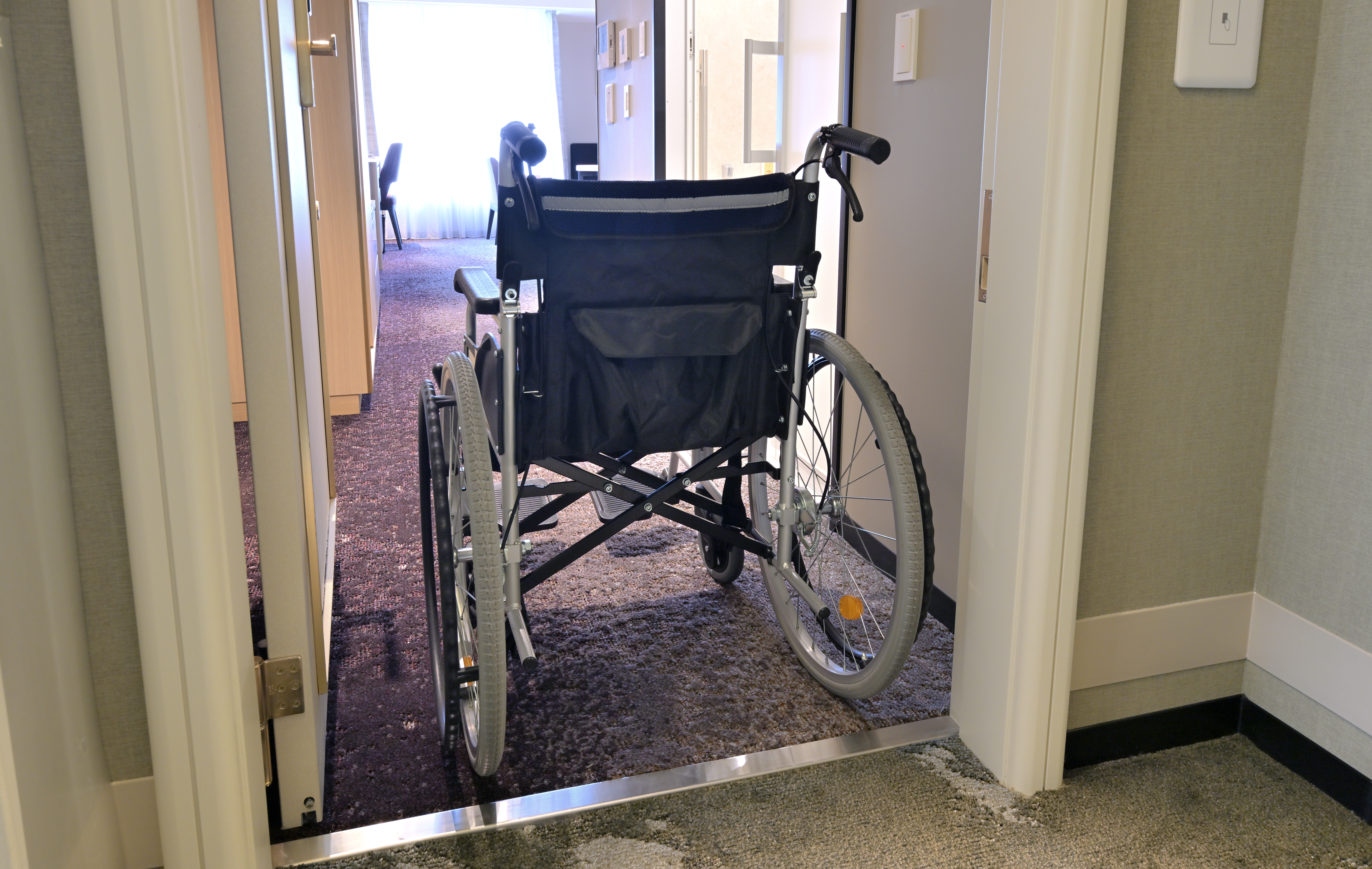 ホテル客室の入口から室内を見た様子。開いたドアの先に手動車椅子が置かれ、車椅子が通れる幅の出入口と、段差の小さい金属製の敷居が見える。奥には客室と明るい窓がある。