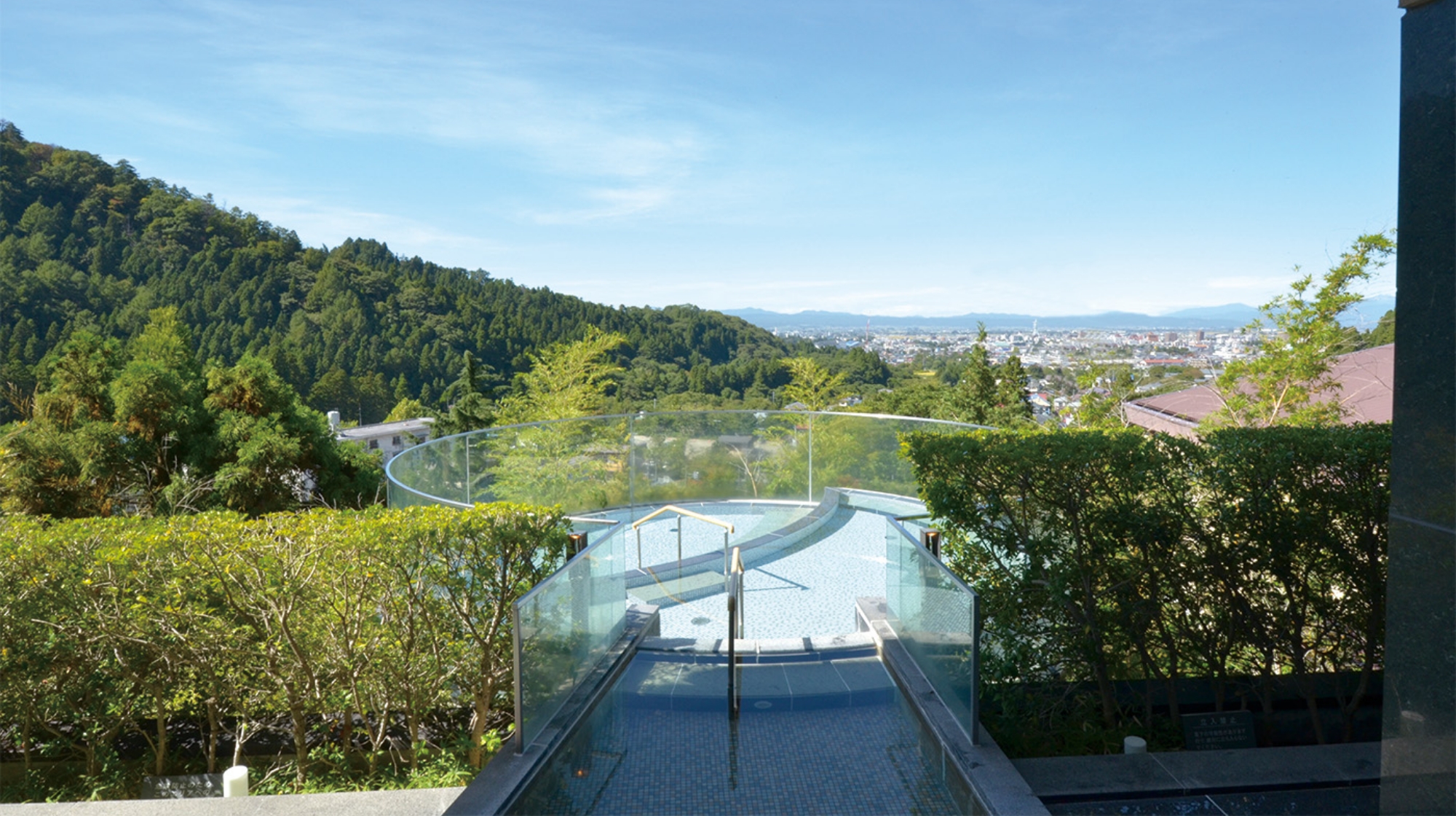 屋外の温泉プール。ガラスの手すり越しに緑豊かな山々と市街地の景色が広がる。晴れた空の下、透明な水がプールに満たされている。