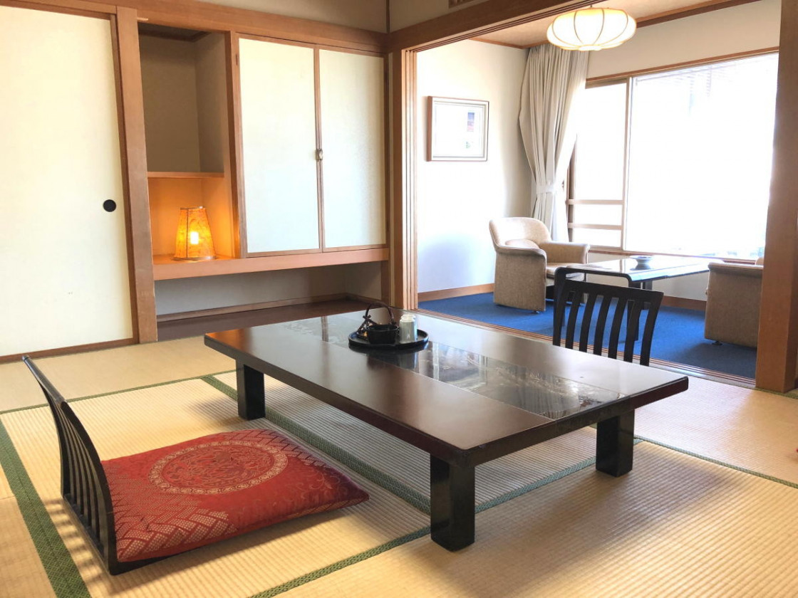 和風の客室である。畳の床に座卓と座椅子が配置されており、隣には障子と床の間がある。窓際には椅子とテーブルが設置されている。