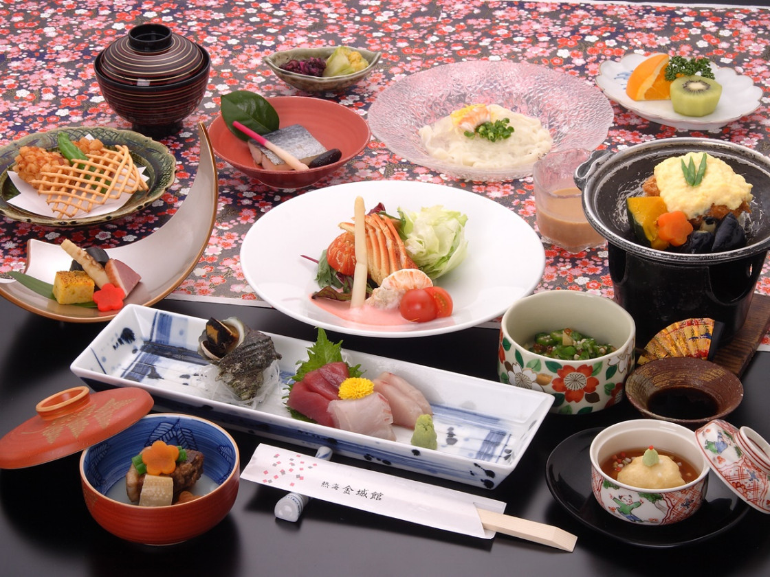 和食のコース料理である。色とりどりの皿に刺身、煮物、焼き物、前菜などが盛り付けられている。テーブルクロスには花柄が施されている。