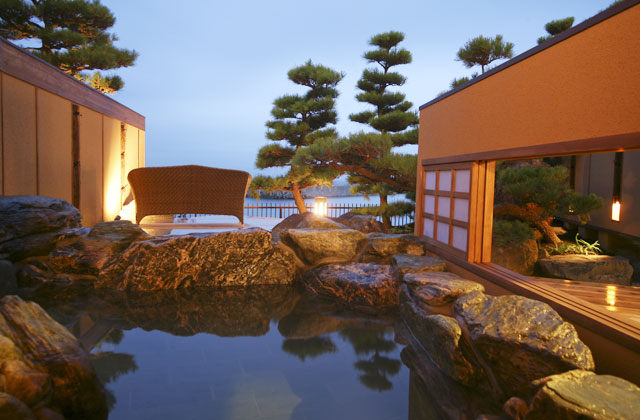 屋外の露天風呂の画像。石造りの浴槽があり、周囲には松の木が植えられている。遠くに海の景色が広がる。