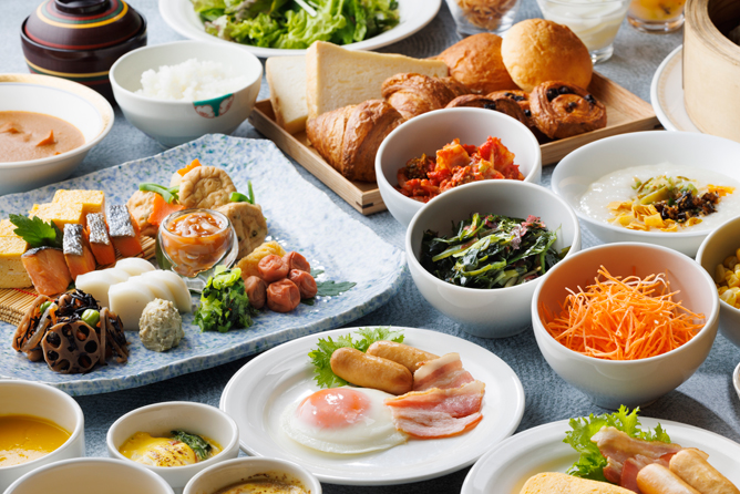 朝食ビュッフェの画像。 様々な料理が並べられている。和食と洋食が両方あり、サラダ、パン、卵料理、小鉢に入った和食の惣菜などが見える。食器は白い皿や小鉢が使用されている。