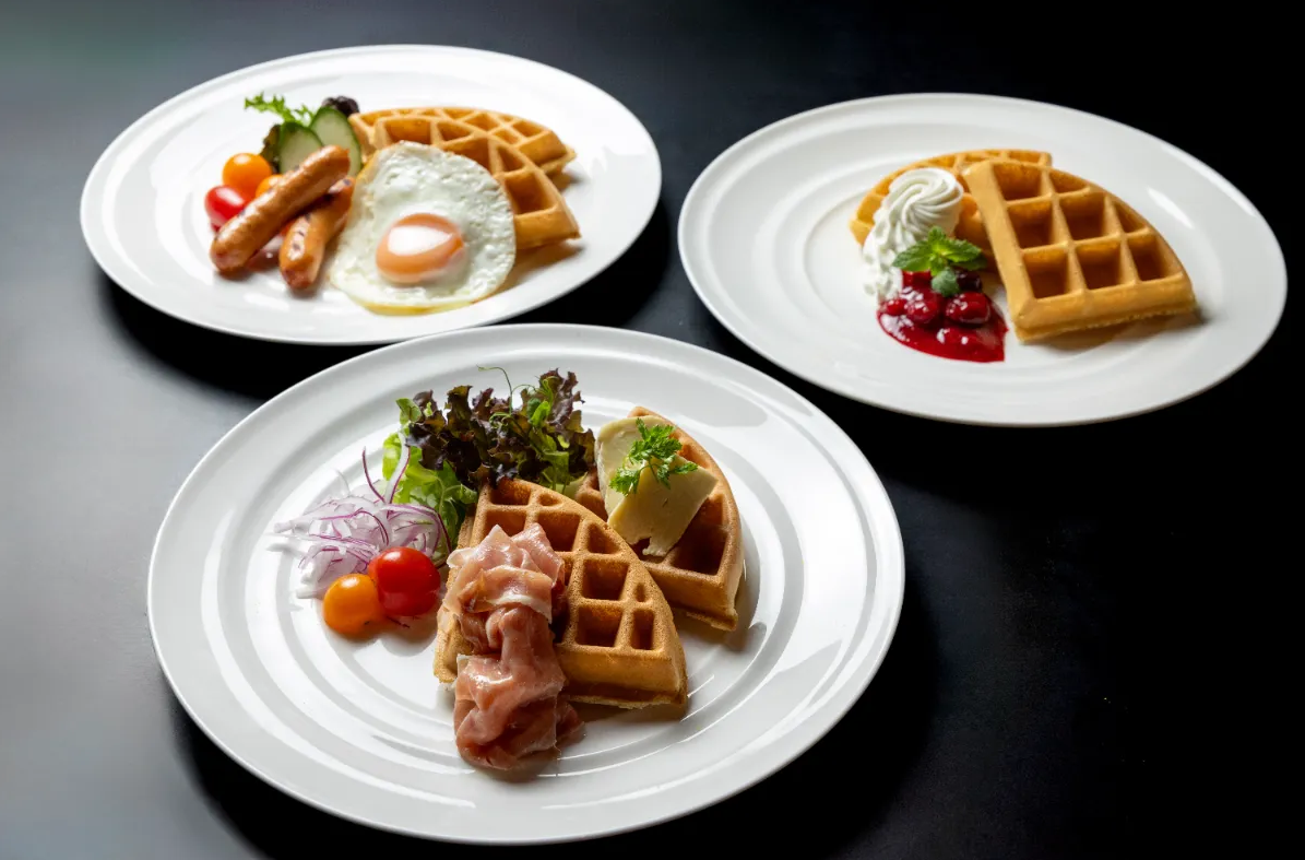 ワッフル料理の画像。 3枚の白い皿にそれぞれ異なるワッフル料理が盛り付けられている。一皿目はソーセージと目玉焼き、二皿目は生クリームとベリーソース、三皿目は生ハムとサラダが添えられている。